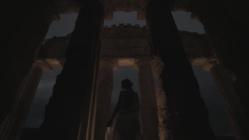 A woman stands beneath ancient columns in the dark
