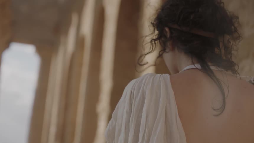 A woman in a pale dress walks near pillars