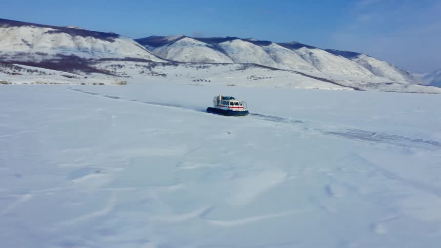 Orbiting aerial view of a hovercraft moving over snowy lake near the mountains