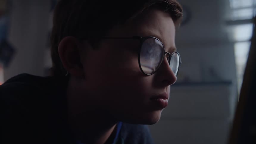 Boy wearing large glasses lays in bed reading storybooks in his bedroom at morning