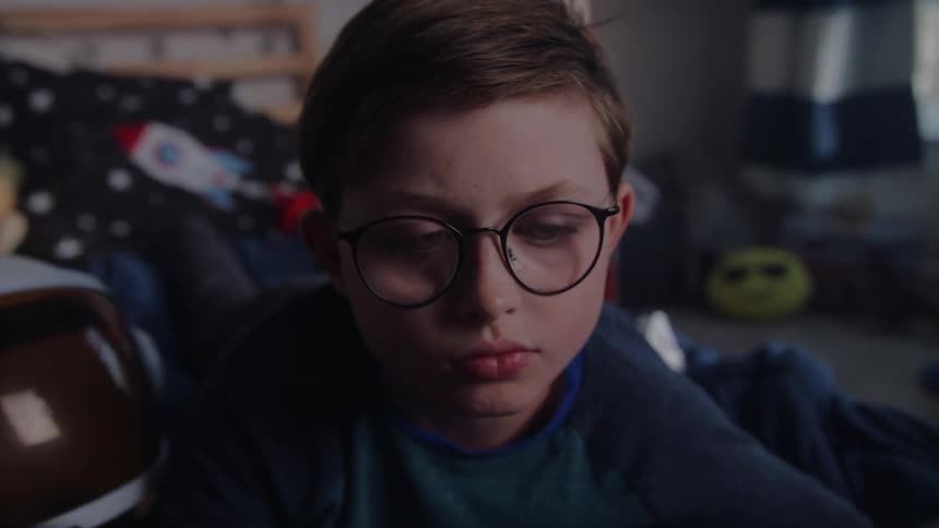 Boy lays in bed reading storybooks near a toy astronaut helmet in his bedroom