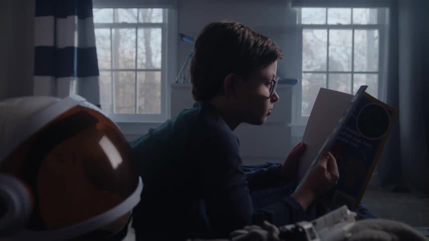 Boy lays in bed reading storybooks near a toy astronaut helmet in his bedroom