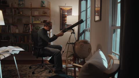 A man adjusts a telescope pointing out of a window of a house