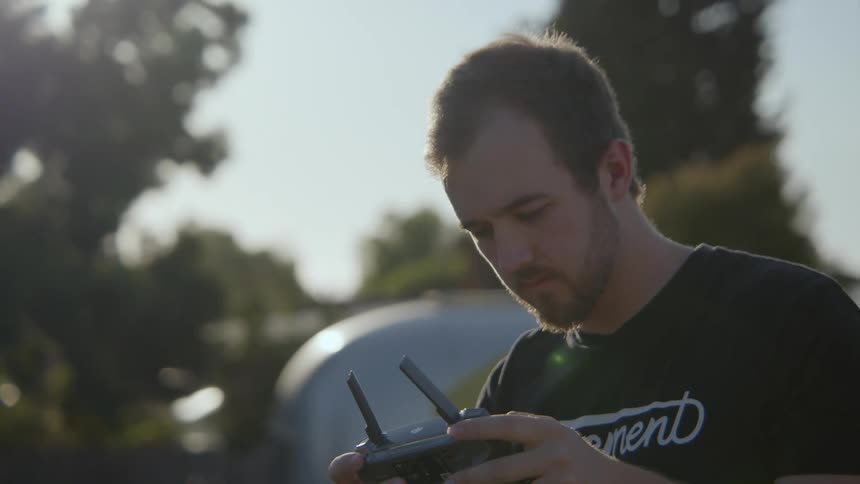 A man standing outside is looking up as he holds a remote controller and he looks back down