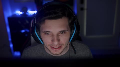 Young male gamer wearing headphones and sitting in front of computer screen and plays video game in dark room smiling and talking
