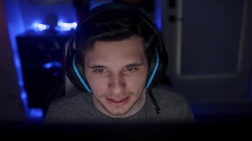 Young male gamer wearing headphones and sitting in front of computer screen and plays video game in dark room smiling and talking