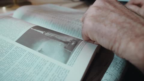 A man is reading a book that is sitting on the table in front of him and he moves his finger across the page
