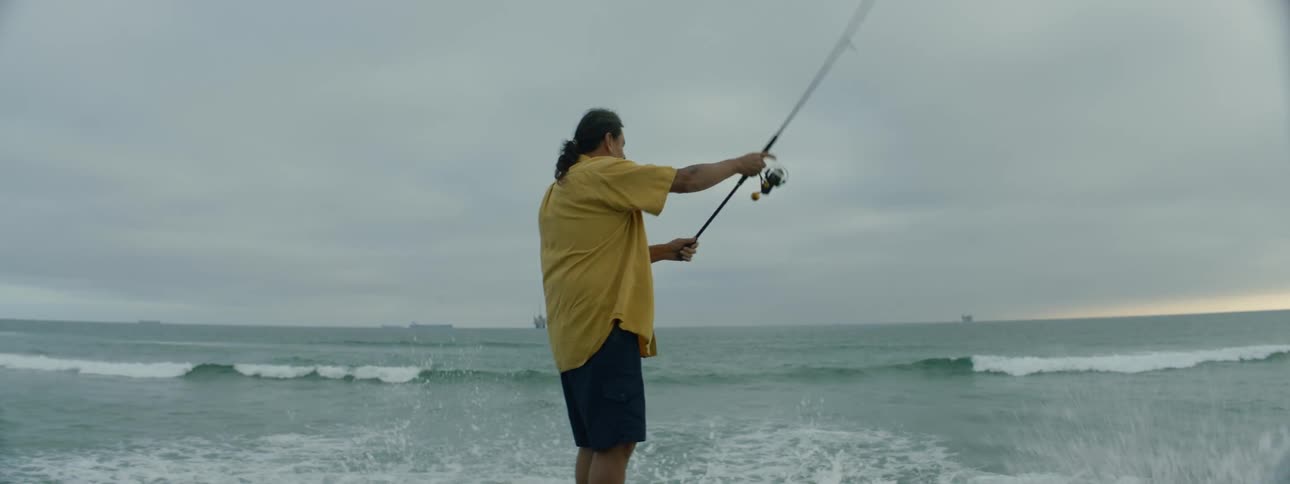 A man is standing along the coast holding a fishing pole and he casts his line as the waves roll into shore