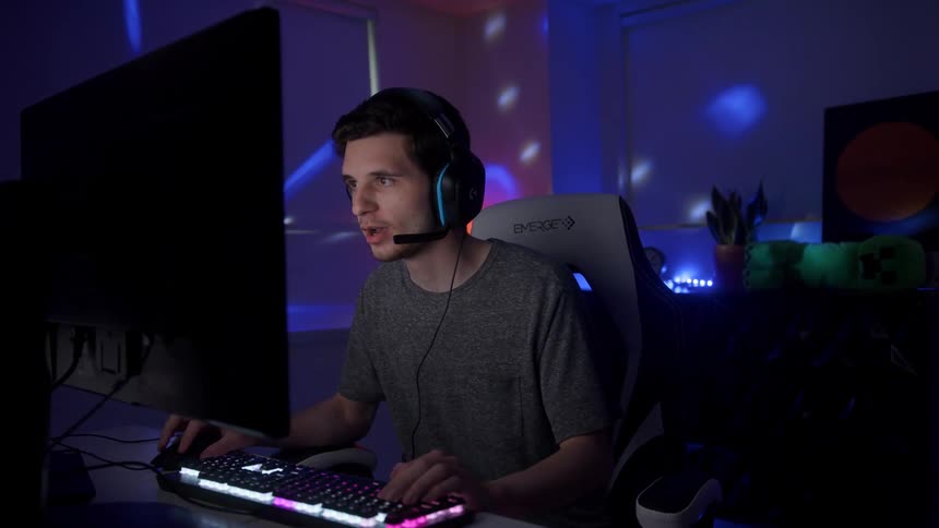 Young male gamer wearing headphones and sitting in front of computer screen and plays video game in dark room