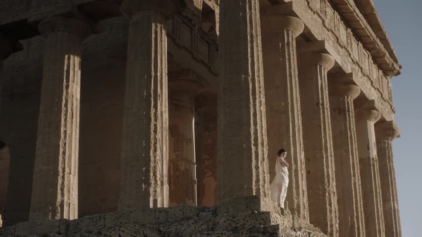 A woman wearing white robes stands by pillars