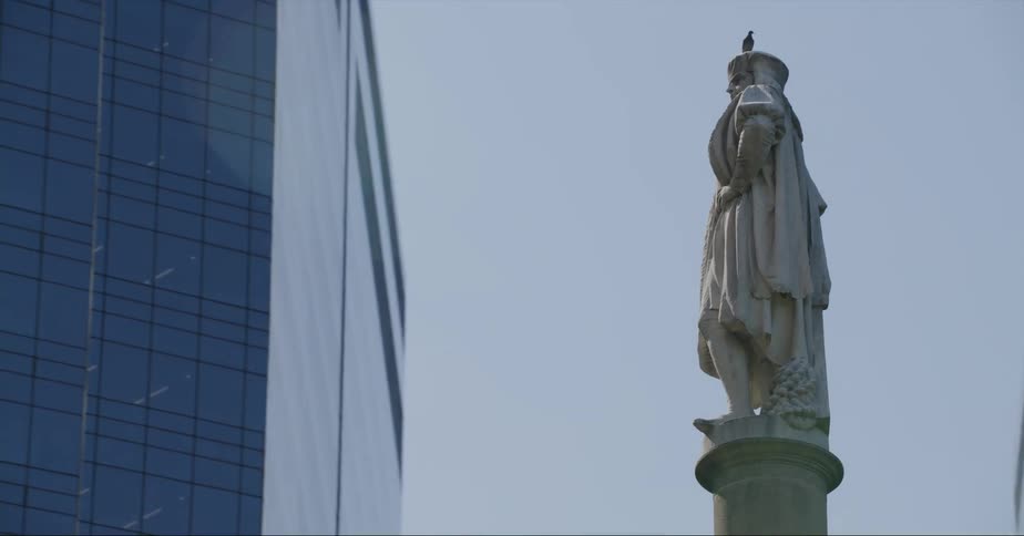 A statue of a man on a pillar surrounded by tall buildings in a city