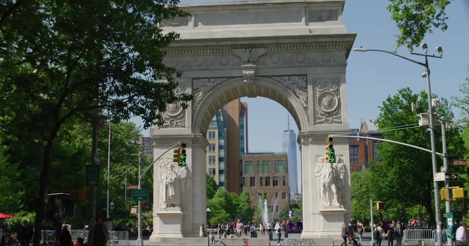 A large group of people walk around a public park by a large arch in a city