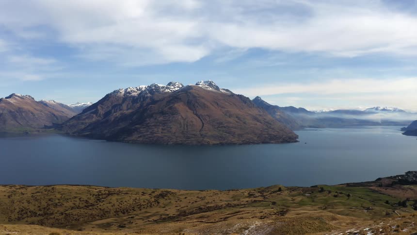 An aerial view of a lake surrounding a mountain range flying over rocks and tussock grass