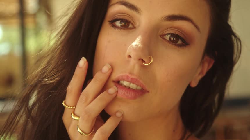 Woman with dark wavy hair rings and nose piercing touches her hair and face looking at camera smiling
