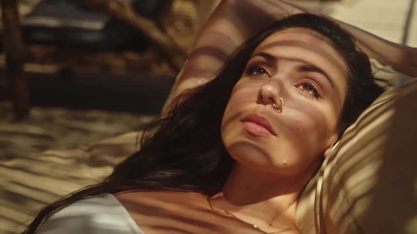 Young woman wearing a white dress reclined on a couch outdoors shields her eyes from the sun