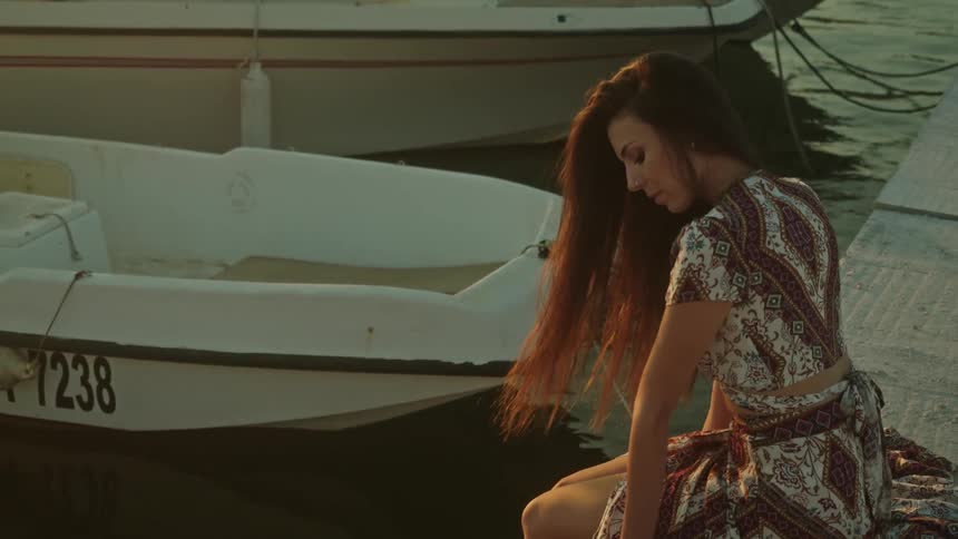 Young woman wearing a long dress sitting on a pier next to small boats at evening