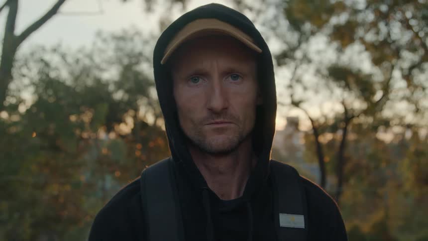 A man stands staring straight ahead with his hood pulled up over his cap