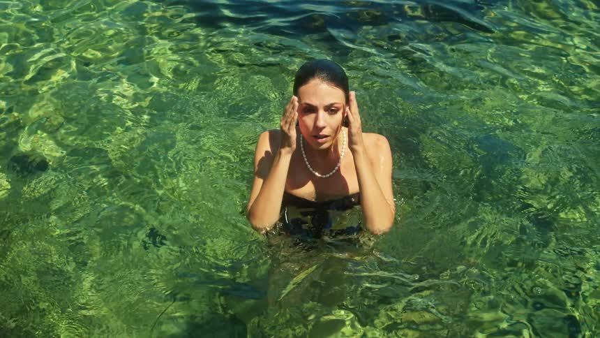 Young beautiful woman wearing a black swimsuit swims out of the water to look at the camera