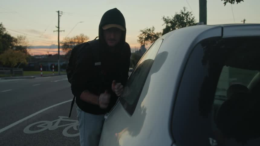 A burglar wearing a hoodie forces the door of a car parked on the street of a town at evening