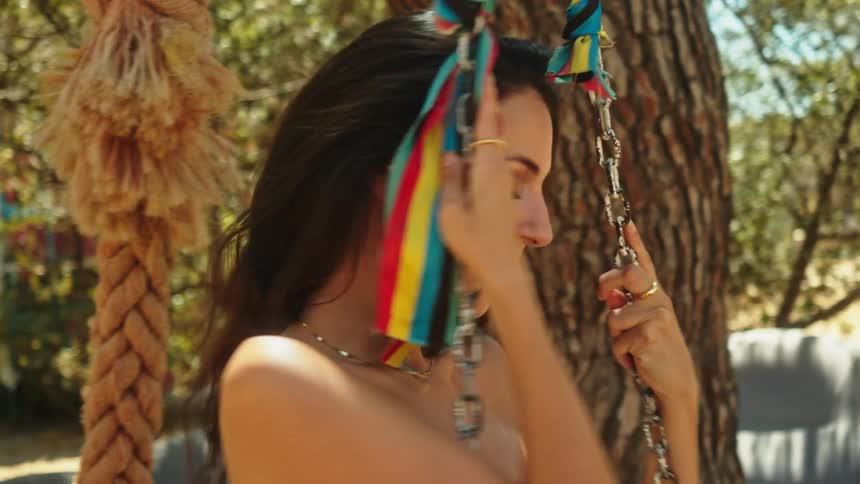 Woman with dark hair necklace and white top swings in swing under the tree smiling
