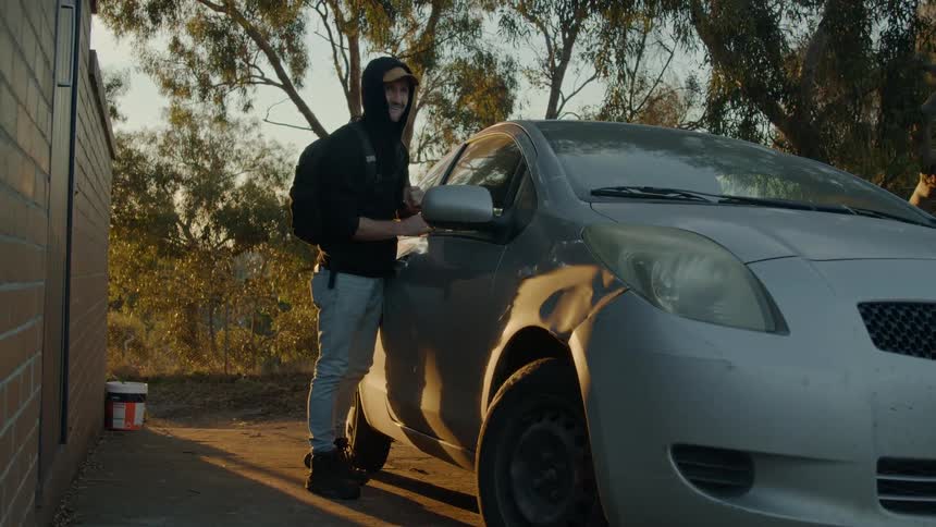Burglar wearing a hoodie forces the door of a car parked near a house at evening
