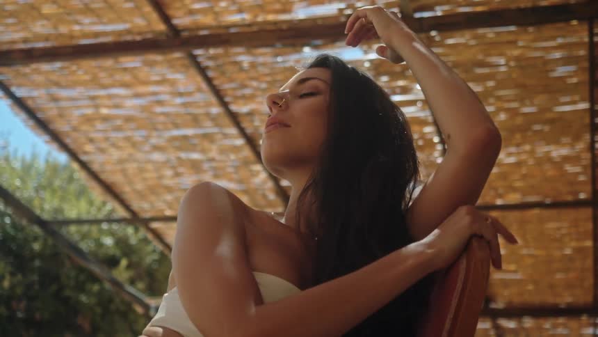 A young woman sits outside under a shade with her eyes closes and stretches her arms and rubs her hair