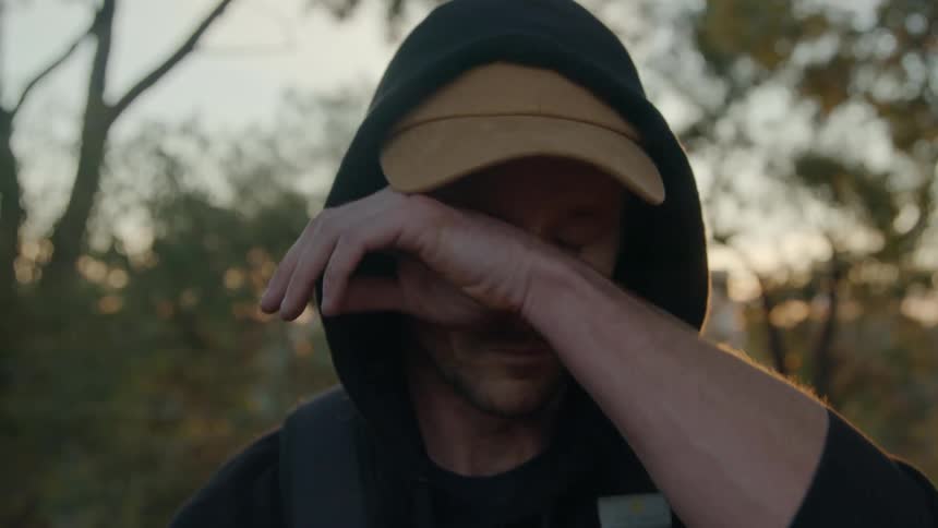 Man with cap black hoodie and backpack stares at camera sniffing at sunset