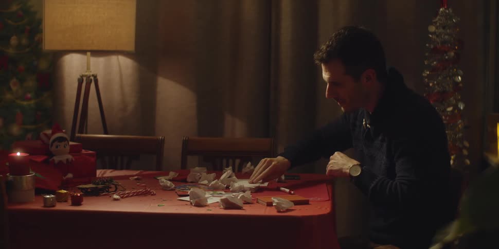 A man sits at home and writes on a piece of paper and then scribbles on it and in frustration throws it on the table where other crumpled up pieces of paper lie