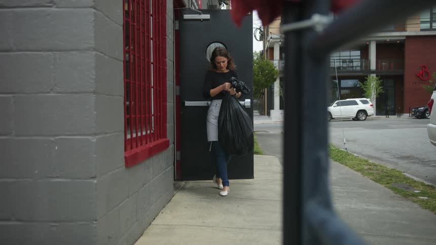 A woman wearing an apron and carrying a bag full of trash opens a door to a building and walks towards the camera