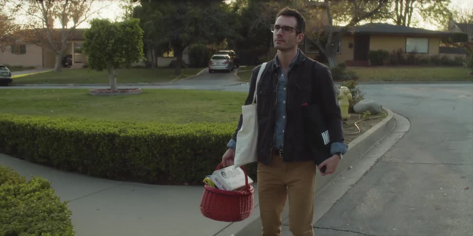 A man walks slowly up a driveway with a basket full of items and a bag under his arm