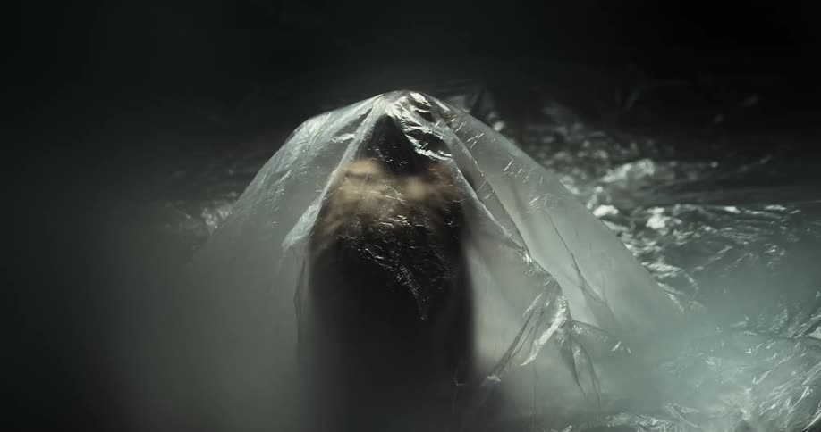 A nude person sits with their back to the camera under a huge sheet of clear plastic and slowly rises to their feet