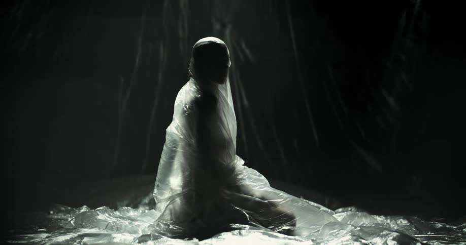 A woman completely wrapped in plastic kneels on the ground and turns her head