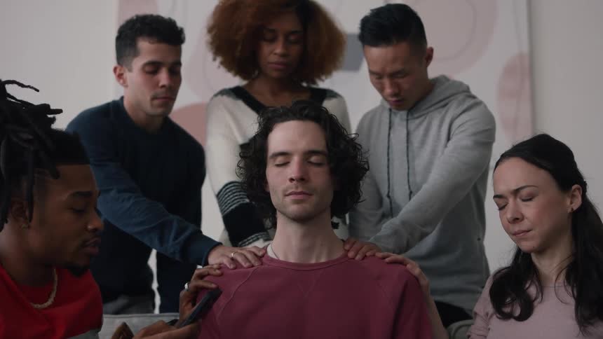 A small group of people are meditating with a hand on one mans shoulders