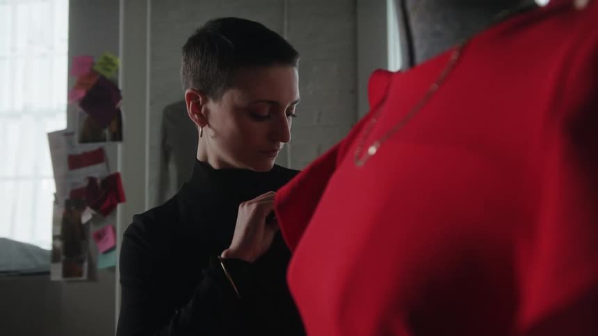 A fashion designer removes a clothing pin from her shirt and uses it to pin clothing on a mannequin