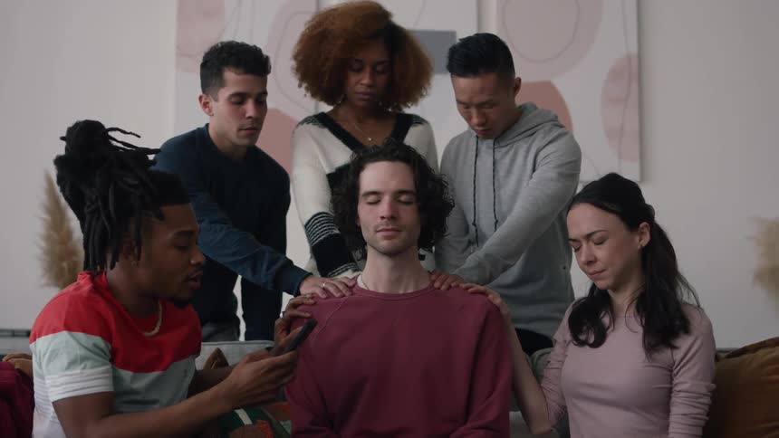 Group of friends rest their hands on the shoulders of a young man sitting on a couch with his eyes closed