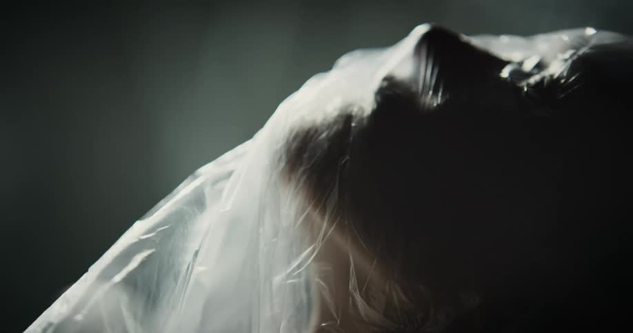 A woman moves her head under a translucent plastic bag in front of a dark background