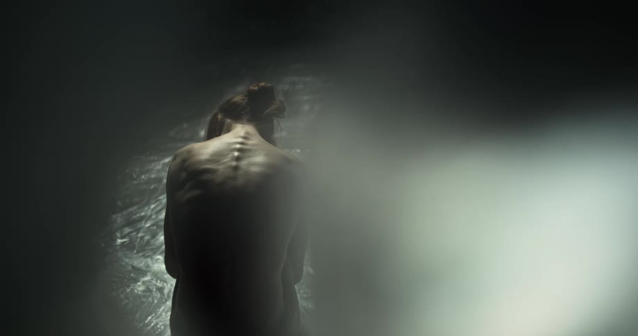 A nude woman is sitting with her back turned and the bones on her spine and ribs stick out as she touches her shoulder and side with her hands