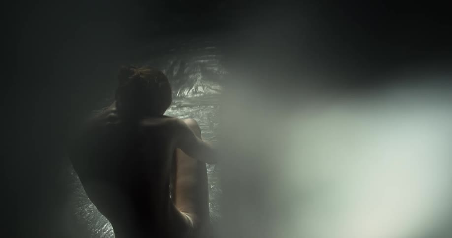 A naked woman sits on the ground surrounded by translucent plastic bags