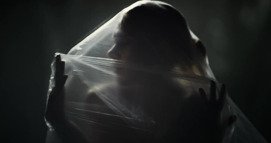 A nude woman is standing with a sheer veil over her body and she uses her hands to push it away from her face as she turns and looks around