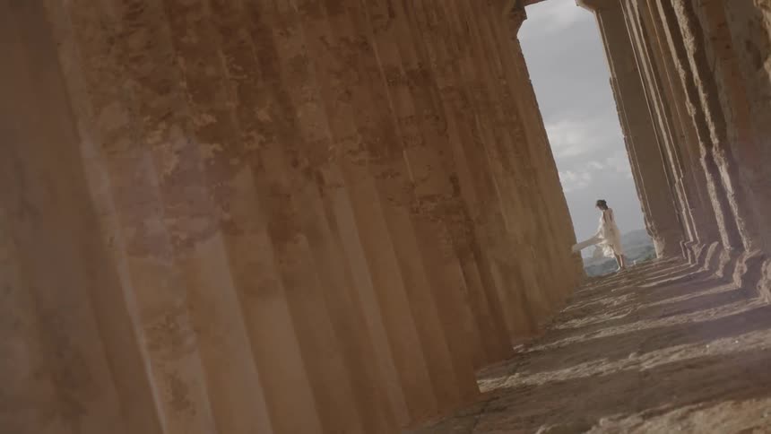 A woman in a flowy dress stands at the end of some ancient columns