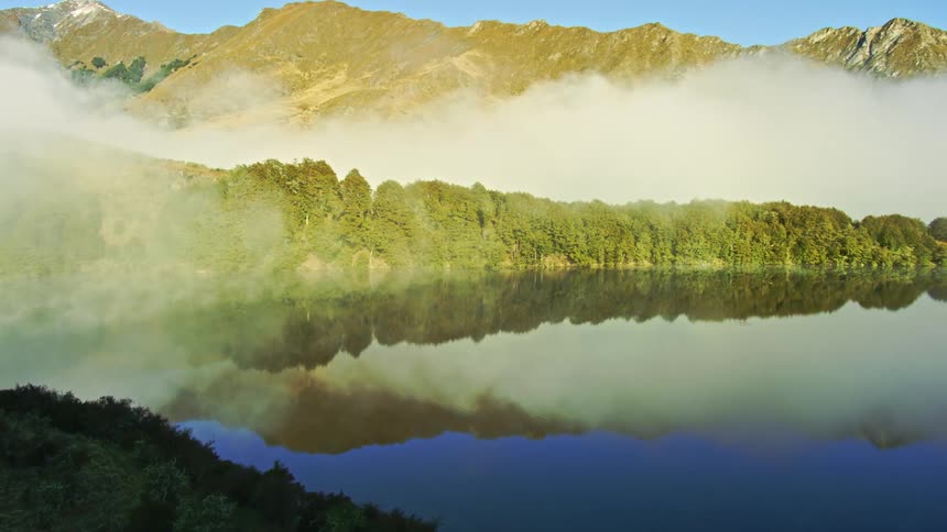 Low white clouds over the slopes of a mountain range next to a reflective lake at morning