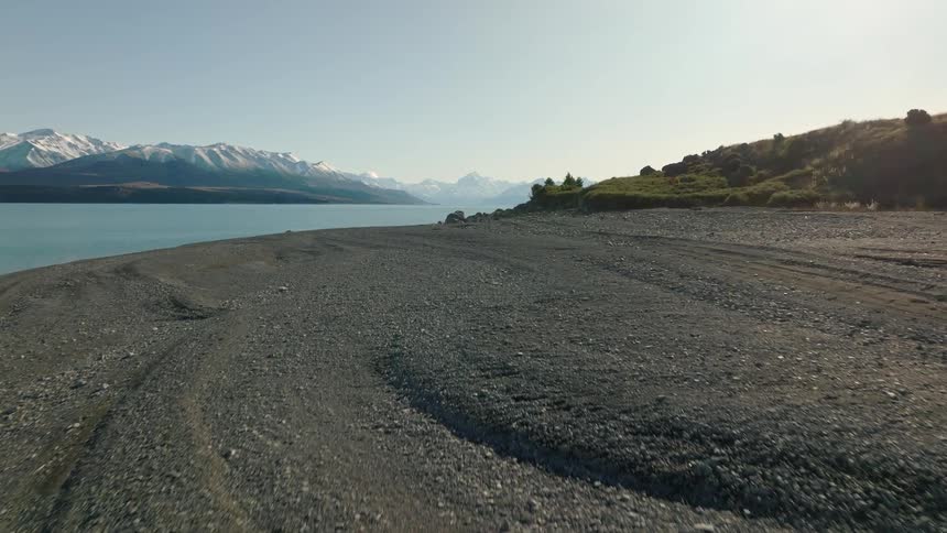 Pebble strewn ground borders hills and water across from snow covered mountains