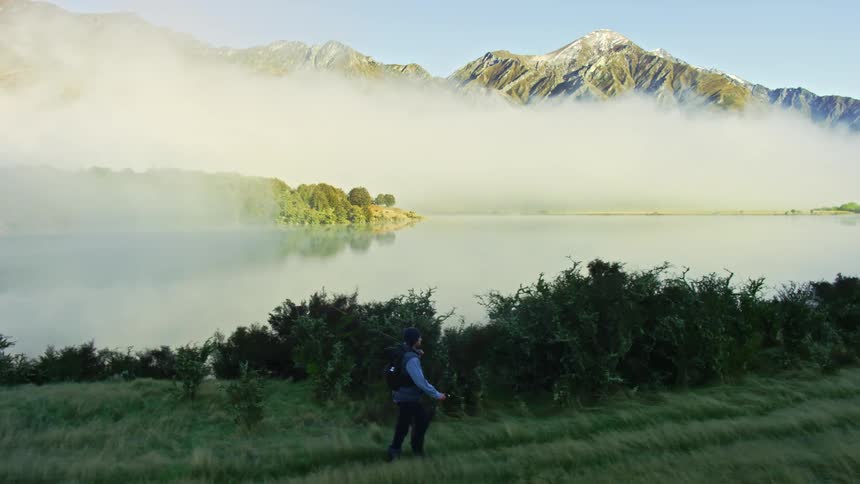 A man carrying fishing gear turns his head to look at the clouds hanging over the lake surface