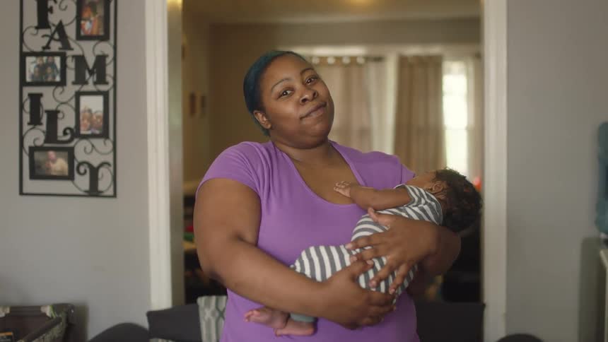 Woman smiles slightly while standing in home holding sleeping baby