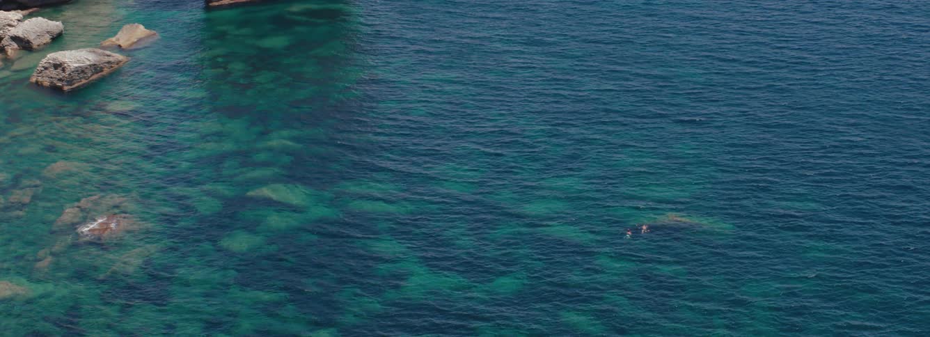Aerial view of two people swimming on the deep blue ocean water close to the coast, in the mediterranean