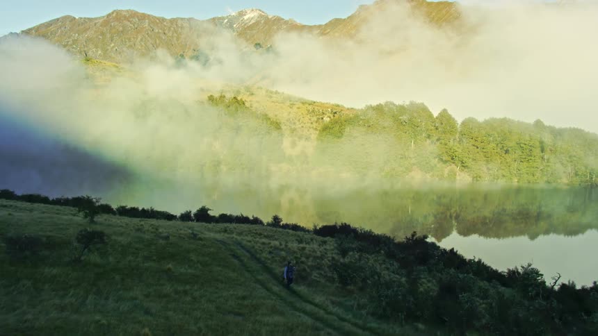 A person walks on a hill next to a lake on a mountain range covered with low white clouds at morning