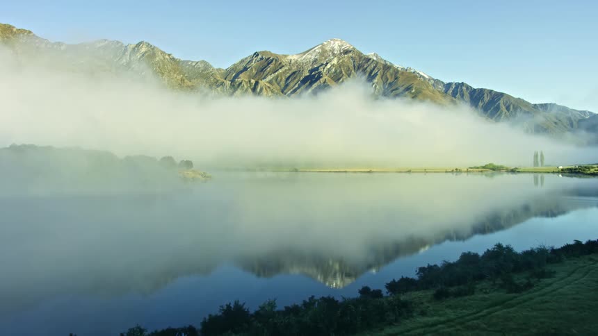 Fog rising on the side of a mountain reflects in the water as trees line the lake under a clear sky