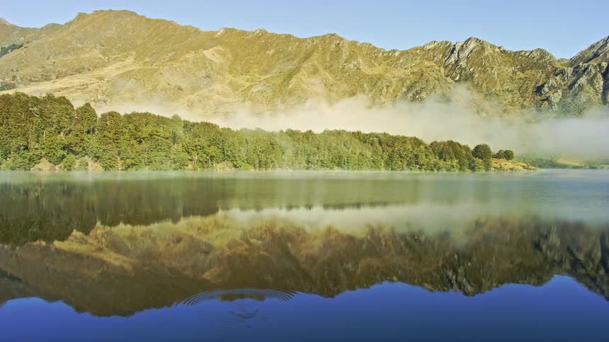 A man is fishing at an alpine lake with reflections and low cloud