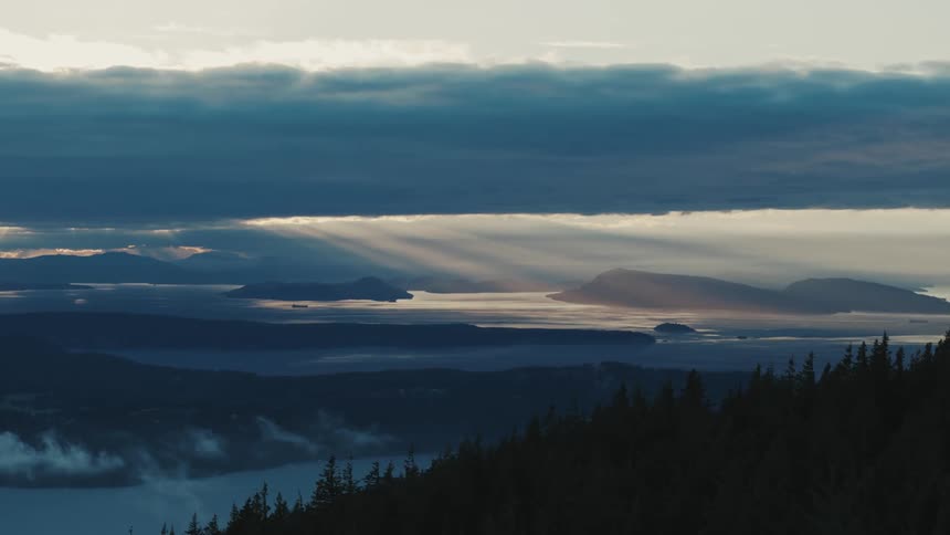 A forest of trees is along the coast and rays of sun shine through low hanging clouds upon the water near the promontory