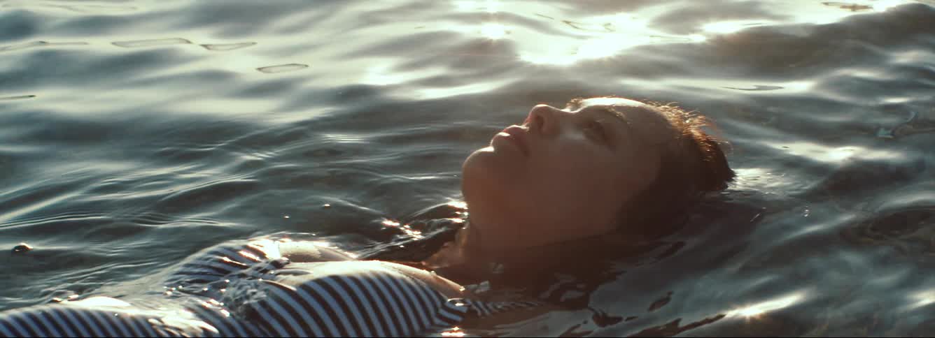A woman in a striped swimsuit floats face up in a body of water during the day, with anamorphic lens flare